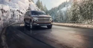 A brown Chevrolet Tahoe SUV driving on a winding mountain road with snowy cliffs and pine trees in the background. | mrchauffeurcolorado