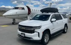White SUV with roof box parked in front of a private jet on a sunny day at the airport.