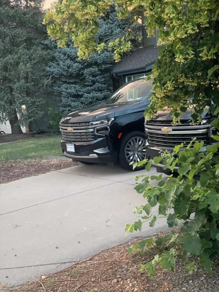 Two black SUVs parked in a driveway near a private house, partially hidden by green trees.