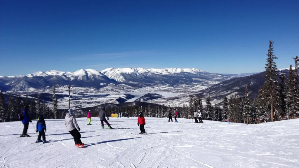 Group of skiers and snowboarders on a snowy mountain slope with panoramic views of snow-covered peaks and a clear blue sky