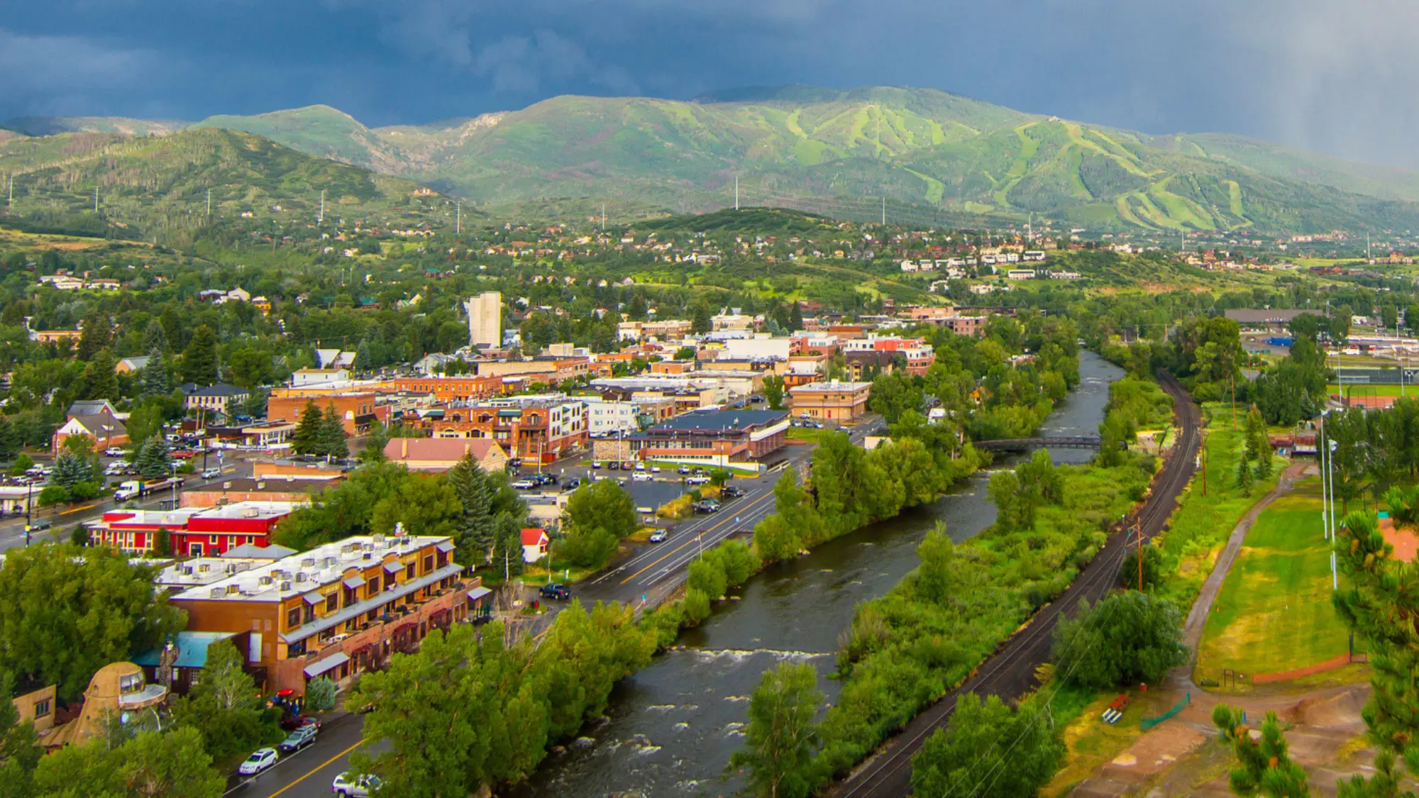 Aerial view of Steamboat Springs in Colorado with a river running beside the main street, a railroad line and green, tree-covered hills with ski runs on the slopes under a moody sky