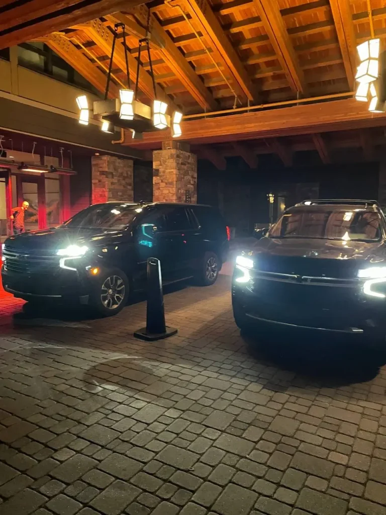Two black luxury SUVs with headlights on parked under a wooden hotel entrance canopy at night