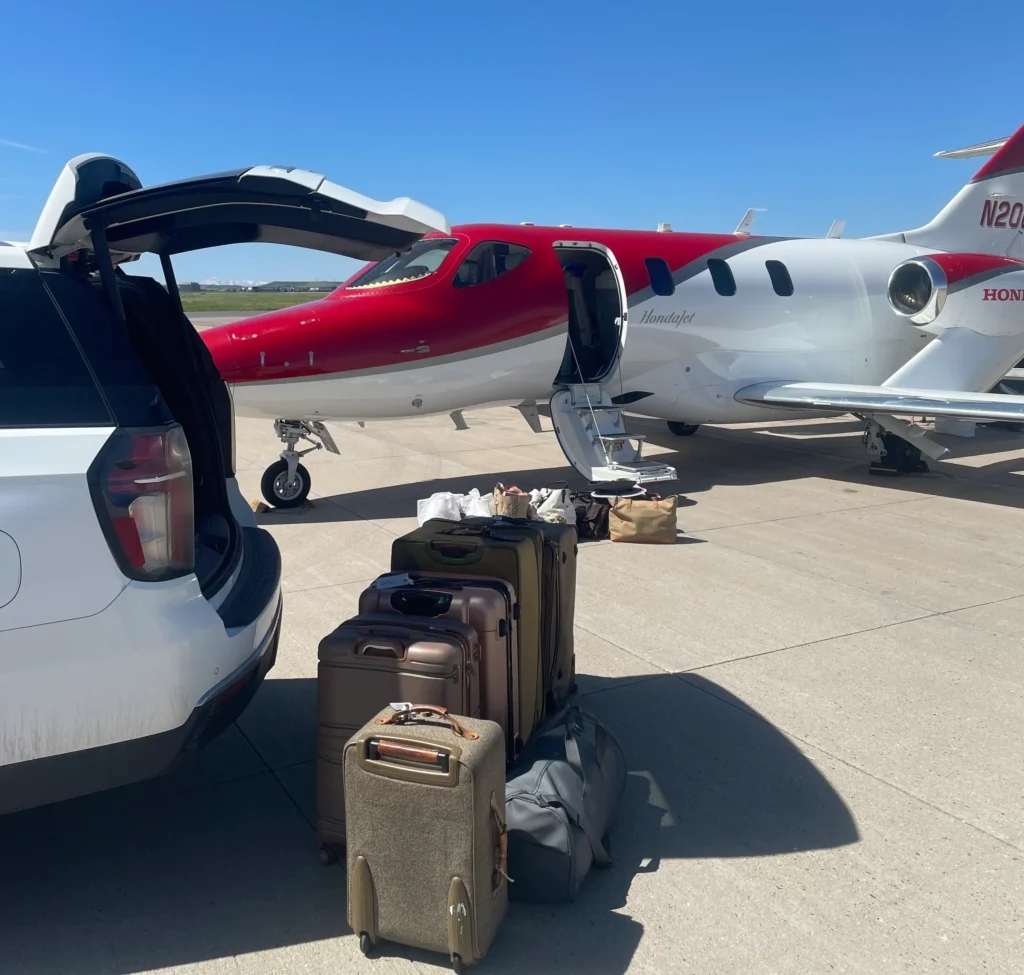 SUV with open trunk and stacked luggage beside a private jet on the airport tarmac under a clear blue sky, illustrating airport transfer and chauffeur service