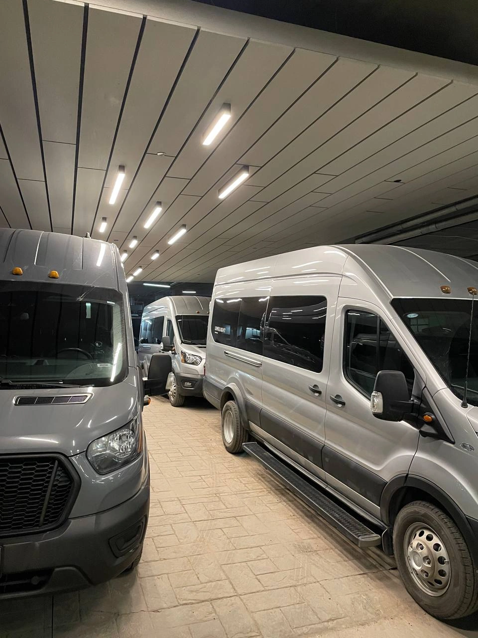 A row of gray passenger vans parked inside a covered garage with overhead lighting.