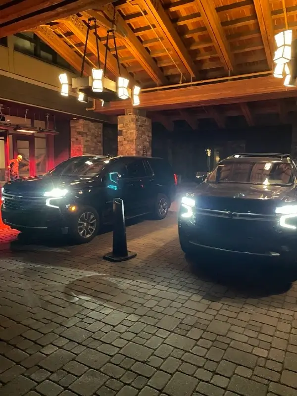 Two black Chevrolet SUVs parked under a covered wooden entrance with stone pillars at night, their headlights illuminated.