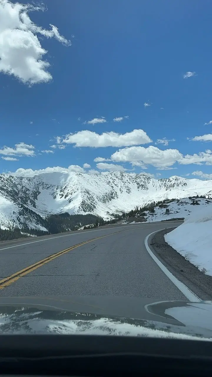 View from a car dashboard looking out at a winding asphalt mountain pass surrounded by deep snowbanks and large, snow-capped mountain peaks under a sunny sky.