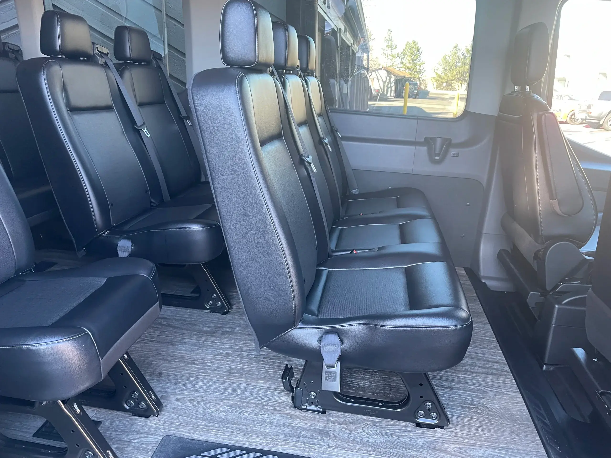 The passenger cabin of a large transit van featuring several rows of black leather seats with integrated seatbelts and a modern grey wood-plank vinyl floor.